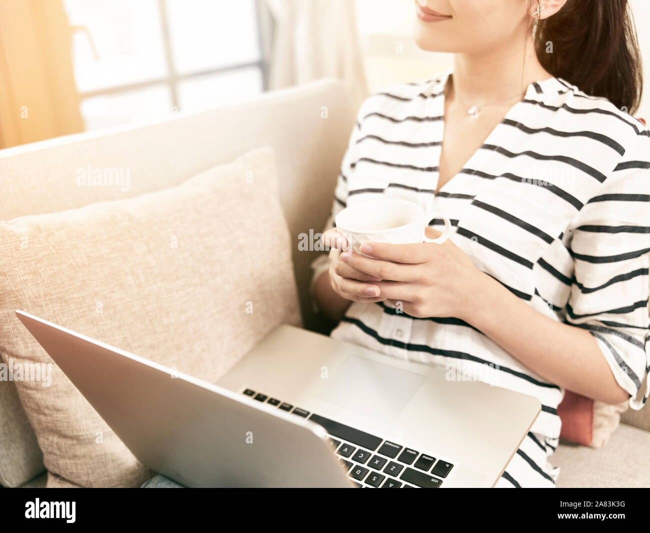 Young Asian woman sitting on couch de boire du café par la fenêtre détendue avec votre ordinateur portable sur les genoux Banque D'Images