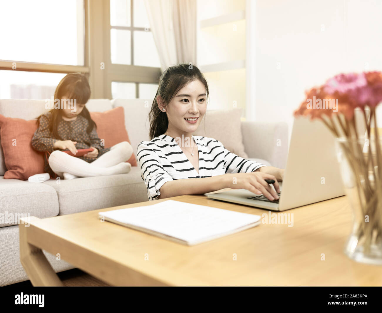 Young Asian woman mère travaillant à domicile à l'aide d'ordinateur portable avec fille en arrière-plan Banque D'Images