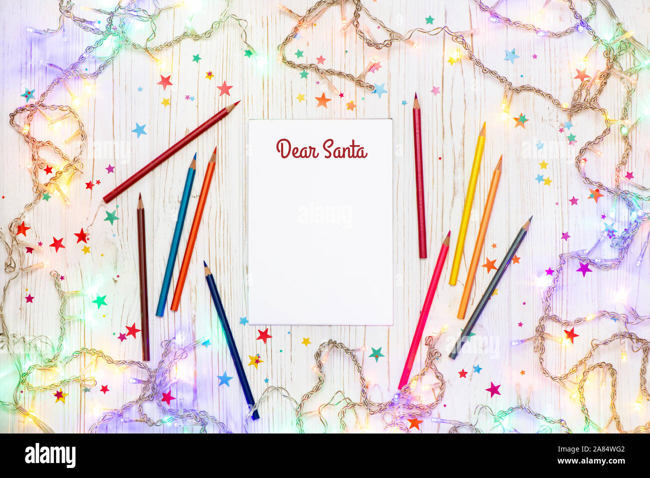 Fond de Noël fête des lumières avec guirlande, colorful crayons et papier blanc avec texte Cher Père Noël sur surface en bois blanc. Banque D'Images