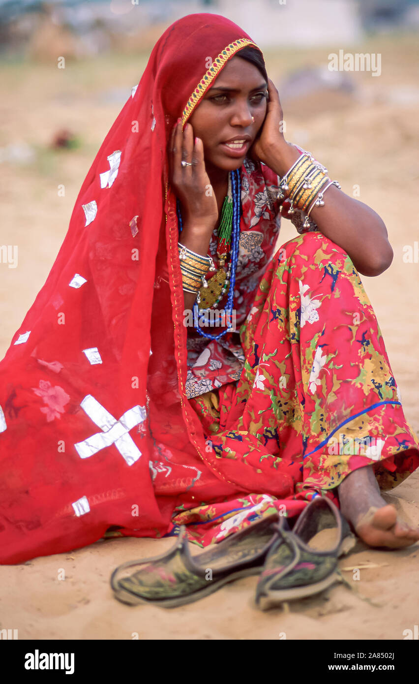 Une belle femme indienne Bhopa assis sur le sable porte un foulard rouge et une jupe multicolore avec de nombreux bracelets et bijoux sur ses bras. Banque D'Images