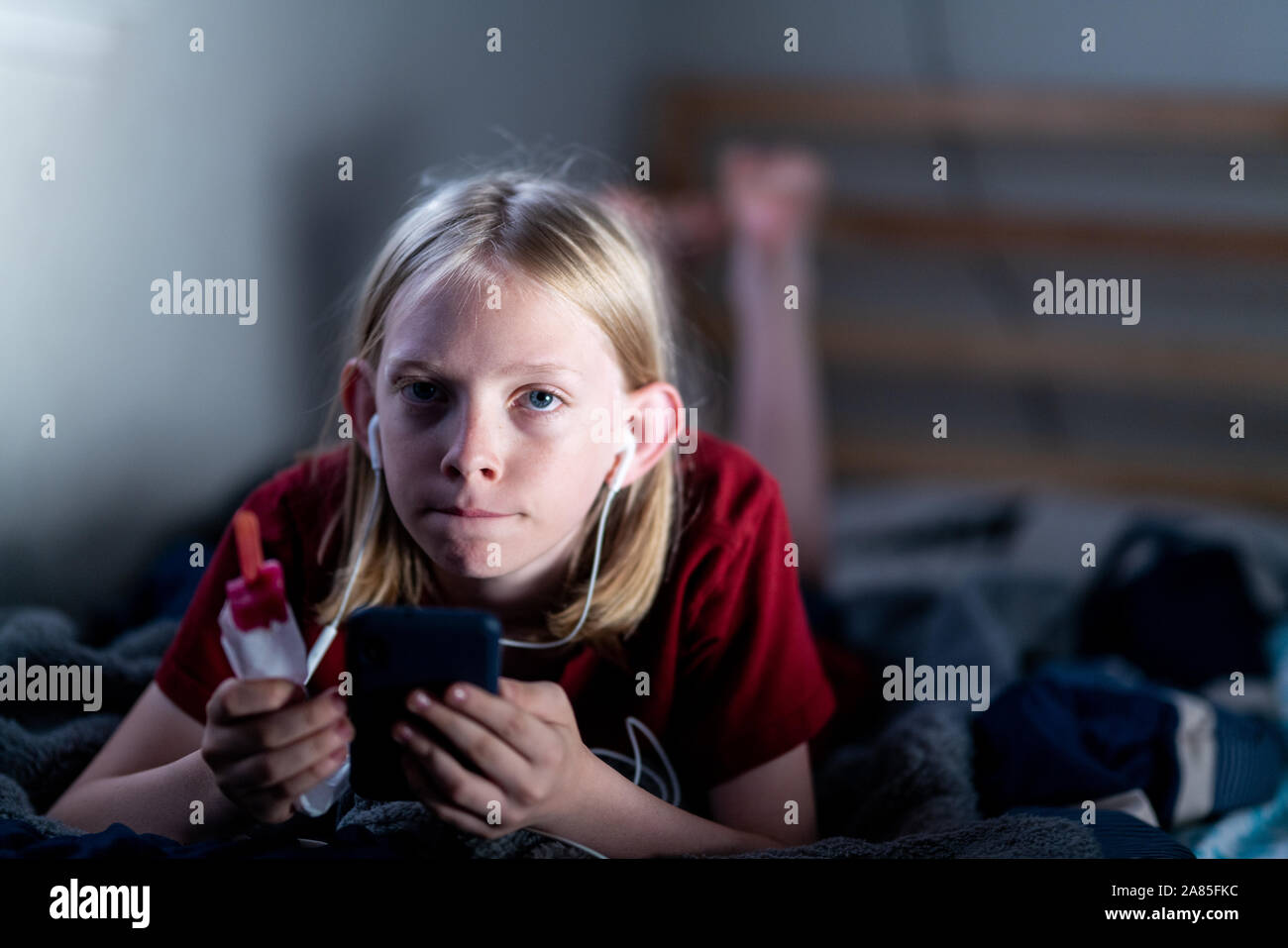 Teenager holding Popsicle lying on bed with phone et écouteurs Banque D'Images