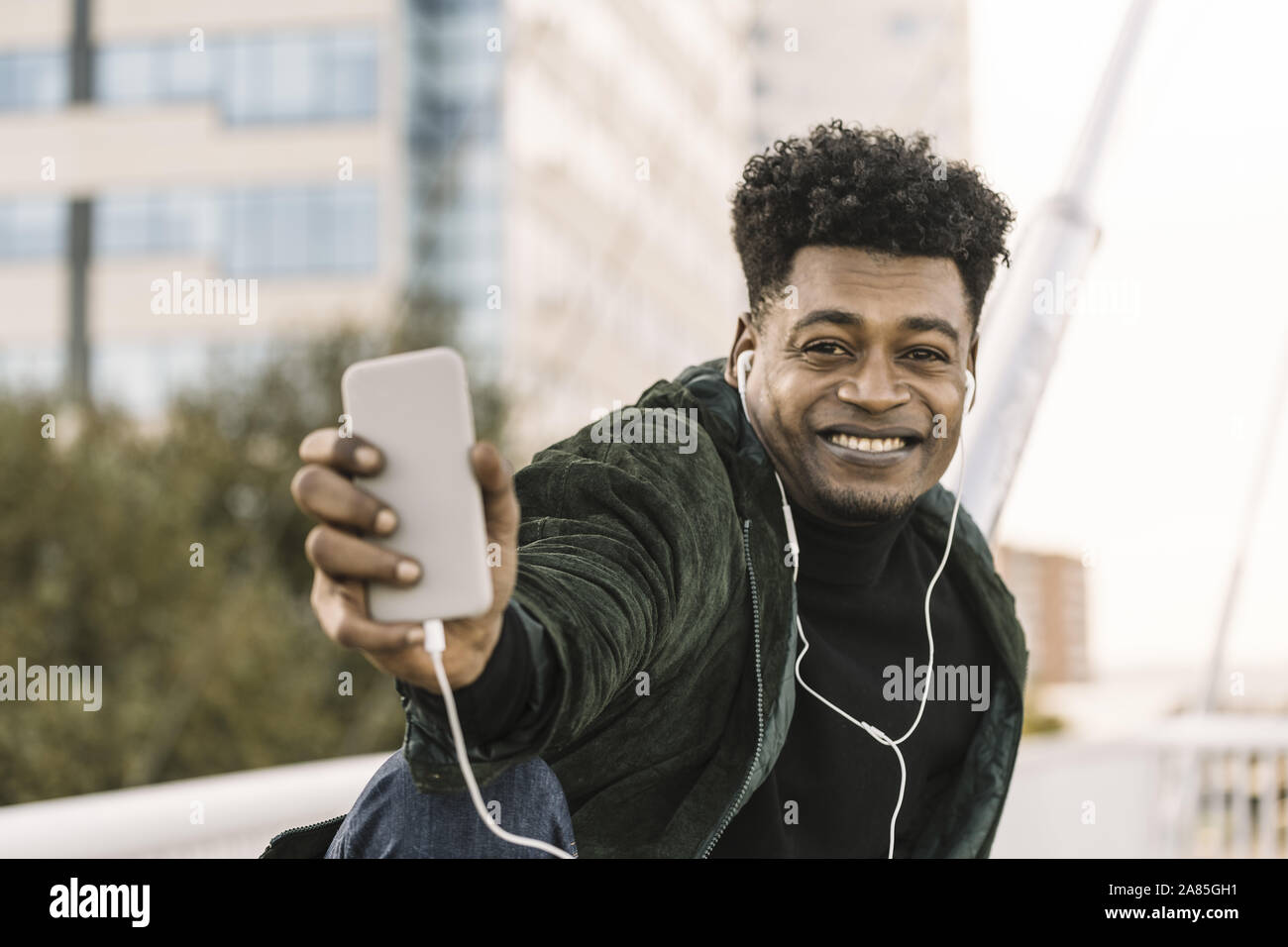 Beau jeune homme noir africain faisant un ou selfies un appel vidéo avec le smartphone et d'écouteurs en position assise, dans la ville, le mode de vie et de techno Banque D'Images