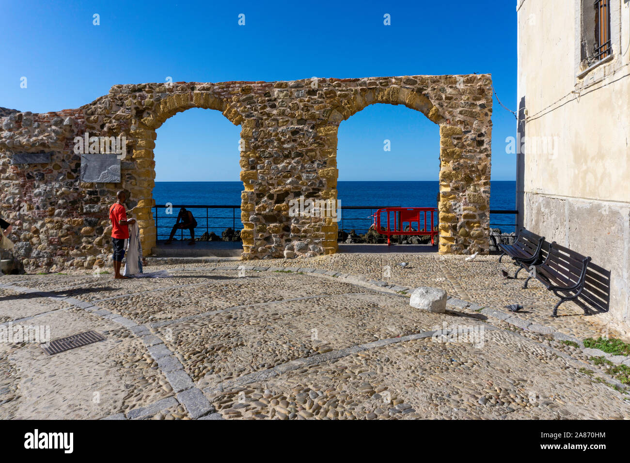 Près de la jetée de Cefalú, Sicile, Italie, un homme debout de vendre ses marchandises en face d'une double arcade menant à la mer. Banque D'Images