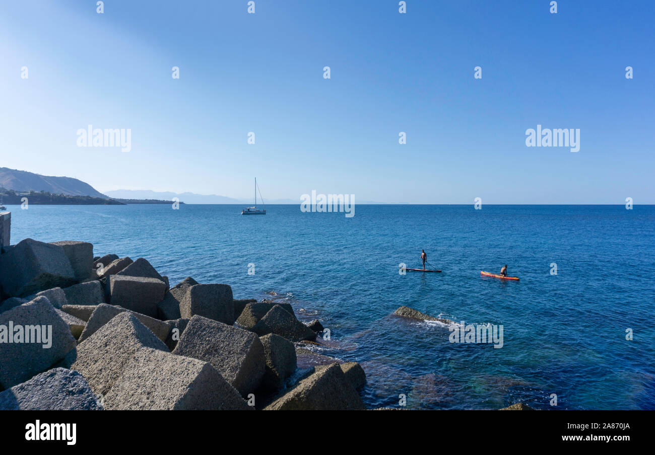 Au large de la côte de Cefalú, Sicile, Italie, deux hommes paddle dans les eaux bleu clair de la méditerranée, avec un yacht au loin. Banque D'Images