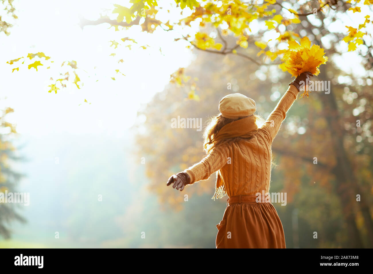 Bonjour l'automne. Vus de derrière, jeune femme insouciante en pull, jupe, chapeau, gants et écharpe avec des feuilles jaunes avec les bras levés à l'extérieur dans la joie Banque D'Images