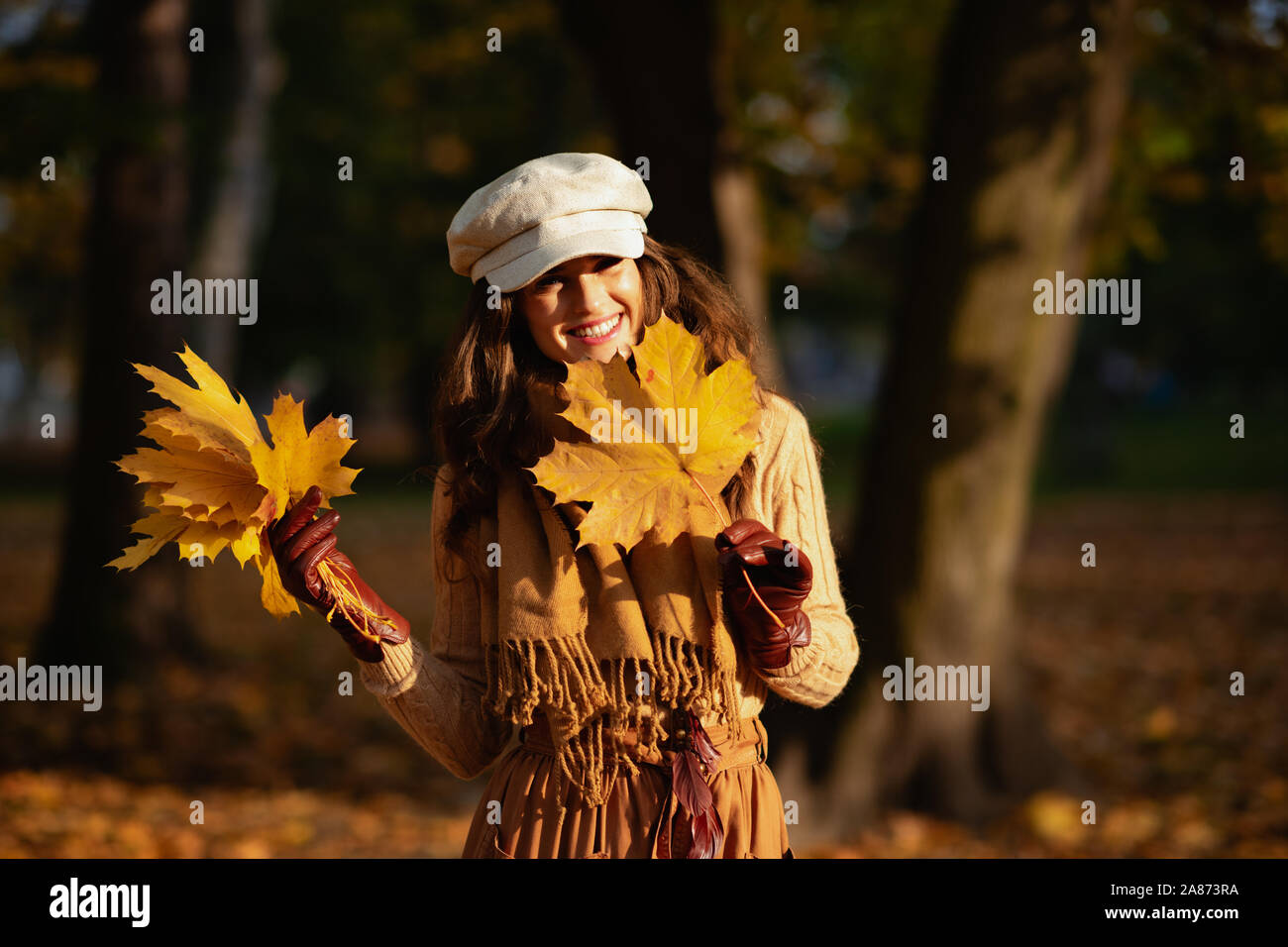 Bonjour l'automne. Portrait of smiling woman in middle age moderne pull, jupe, chapeau, gants et écharpe avec feuilles jaunes à l'extérieur dans le parc de l'automne. Banque D'Images