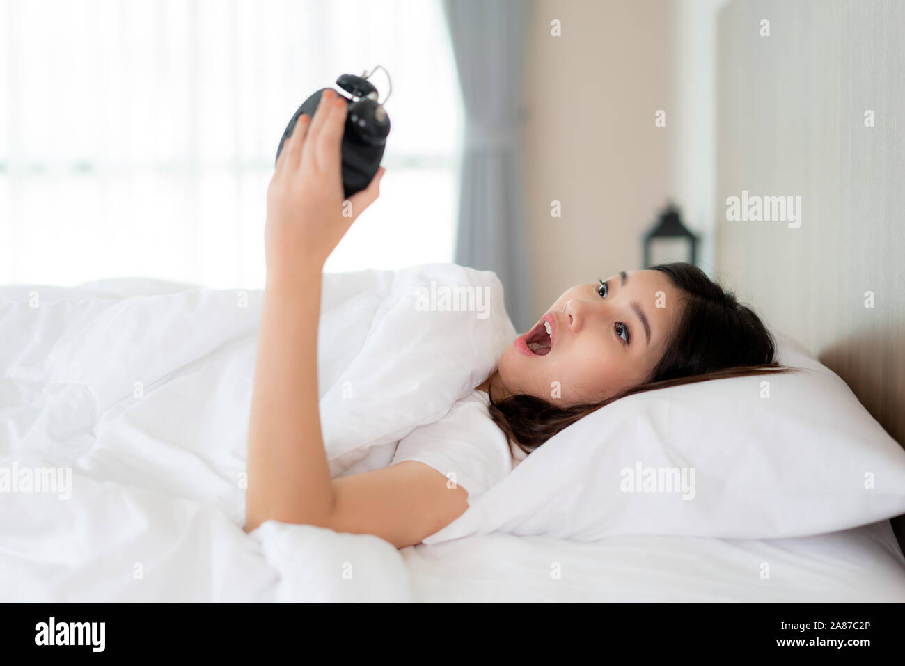 Jolie Asian Woman lying on bed blanc et noir holding réveil avec le choc d'apprendre qu'en fin de matinée à coucher à la maison. Banque D'Images