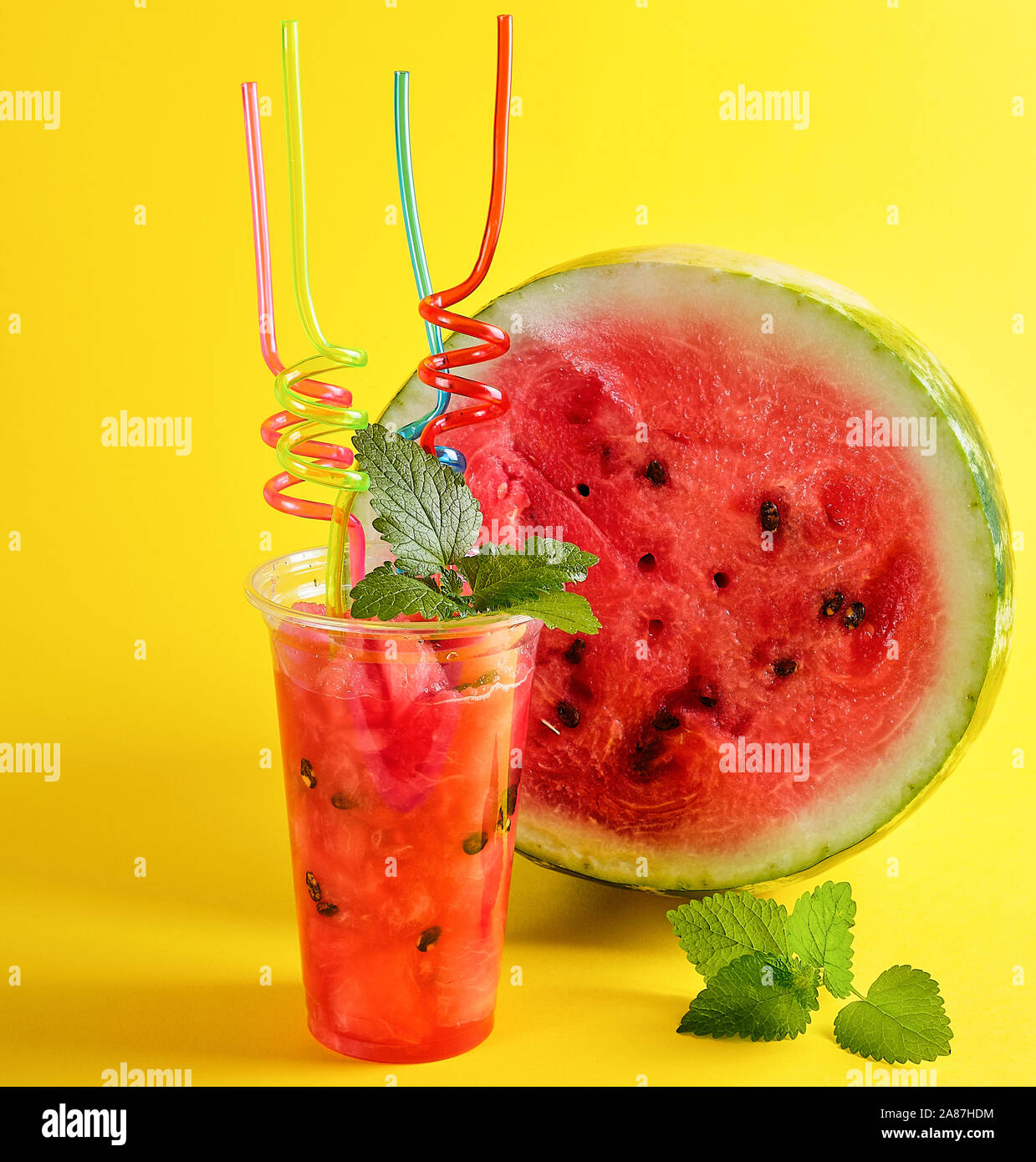 De smoothie pastèque rouge mûr dans une tasse en plastique, lot de tubes colorés et une feuille de menthe sur fond jaune Banque D'Images