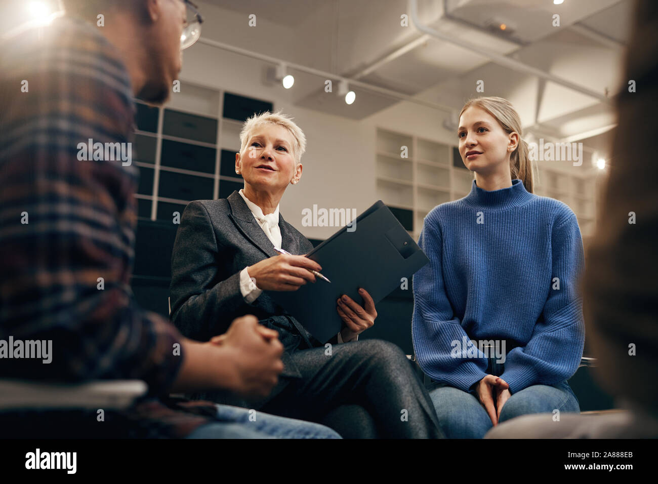 Femme mature avec de courts cheveux blonds holding clipboard et à l'écoute des jeunes racontant leurs plans Banque D'Images