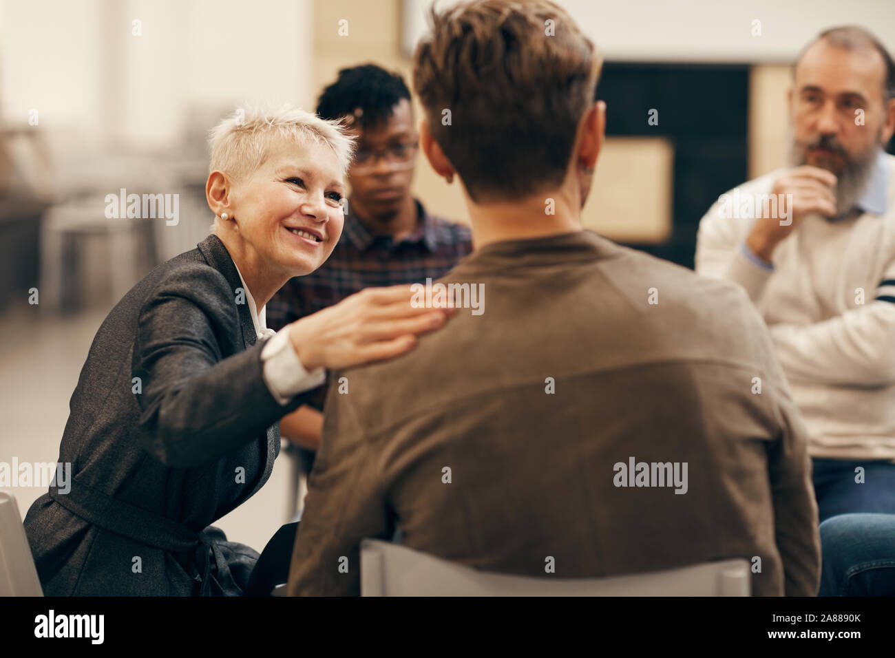 Smiling mature woman avec de courts cheveux blonds de parler au jeune homme qui, assis sur la chaise retour à l'appareil photo pendant la classe thérapeutique Banque D'Images