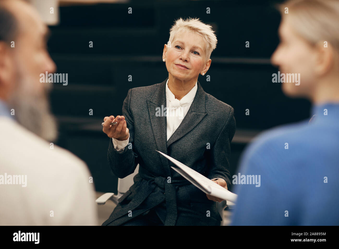 Femme mature avec de courts cheveux blonds holding documents et de parler à ses collègues au cours de réunion Banque D'Images