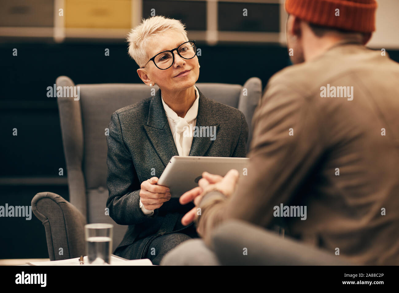 Femme mature avec de courts cheveux blonds sitting on sofa with digital tablet et l'écoute du jeune homme au cours de la réunion d'affaires Banque D'Images