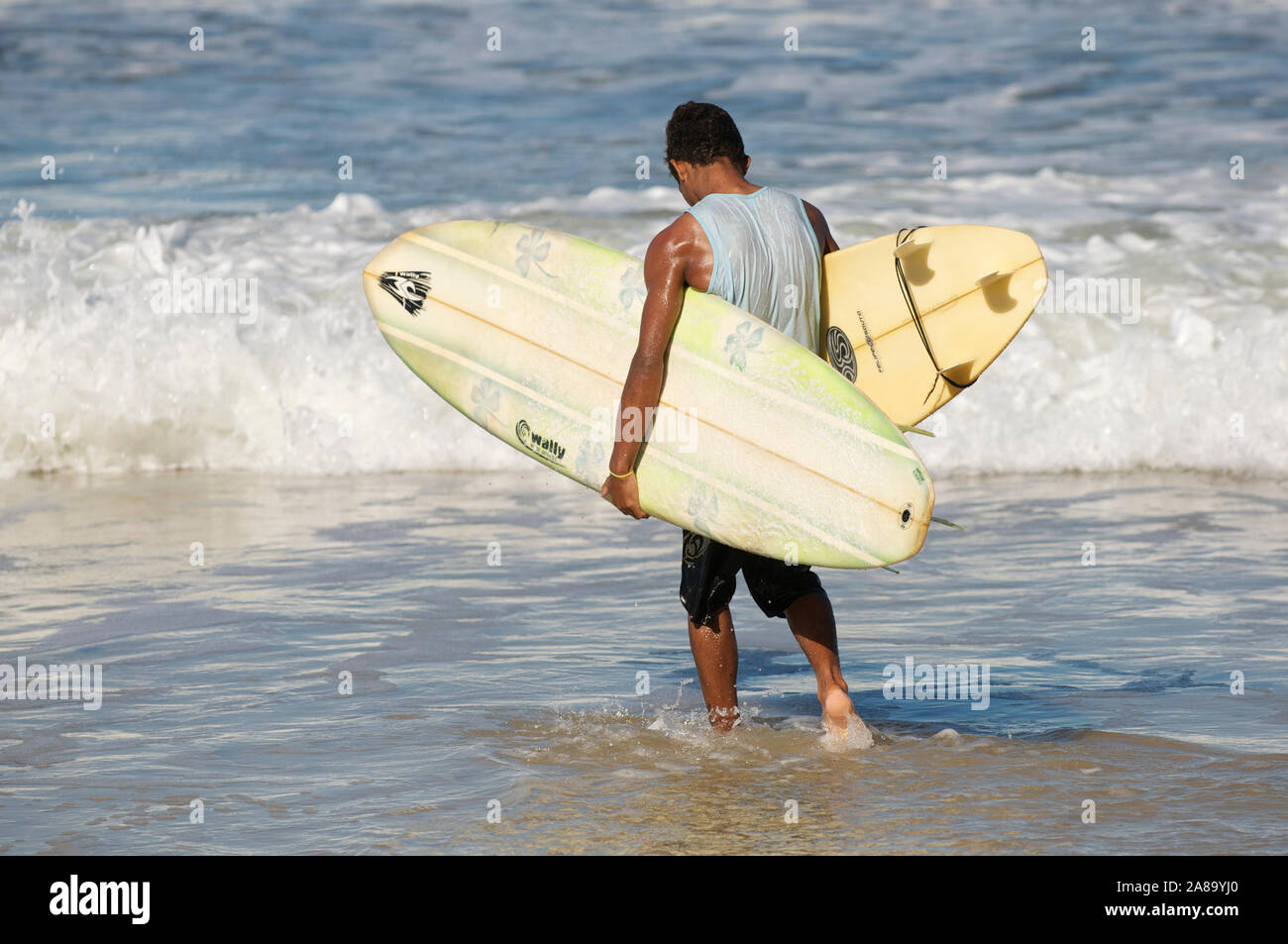 PIPA, RN, BRÉSIL - 6 avril, 2011 : un jeune surfeur brésilien porte deux planches alors qu'il patauge dans les vagues. Banque D'Images