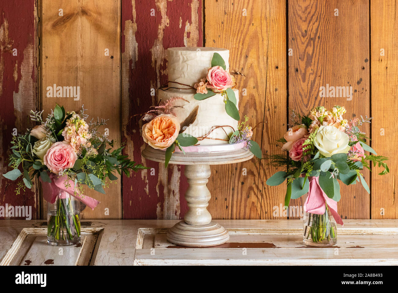 Un gâteau de mariage rustique sur un stand avec des fleurs et des bâtons rétractable le gâteau. Le gâteau se trouve au sommet d'une porte avec du bois grange derrière. Des vases de fleurs sont Banque D'Images