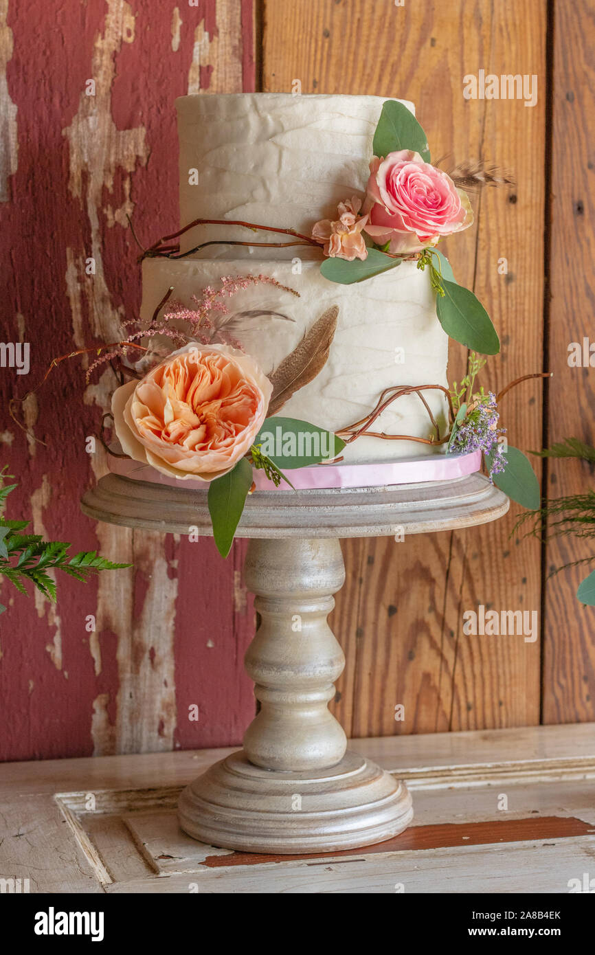 Un gâteau de mariage rustique sur un stand avec des fleurs et des bâtons rétractable le gâteau. Le gâteau se trouve au sommet d'une porte avec du bois grange derrière. Banque D'Images