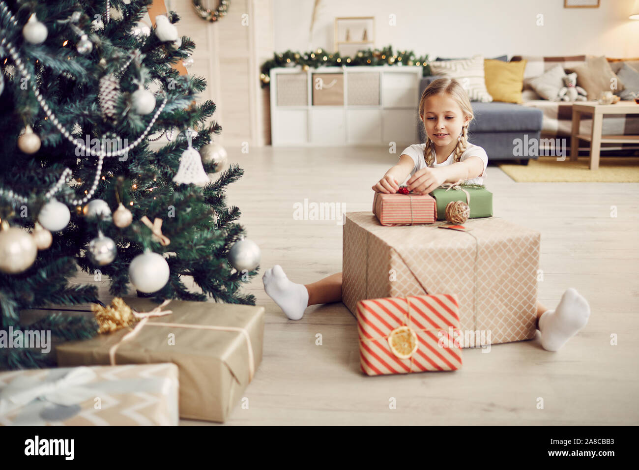 Petite fille aux cheveux blonds assis sur le plancher près de l'arbre de Noël et cadeaux de décoration dans la salle de séjour à la maison Banque D'Images