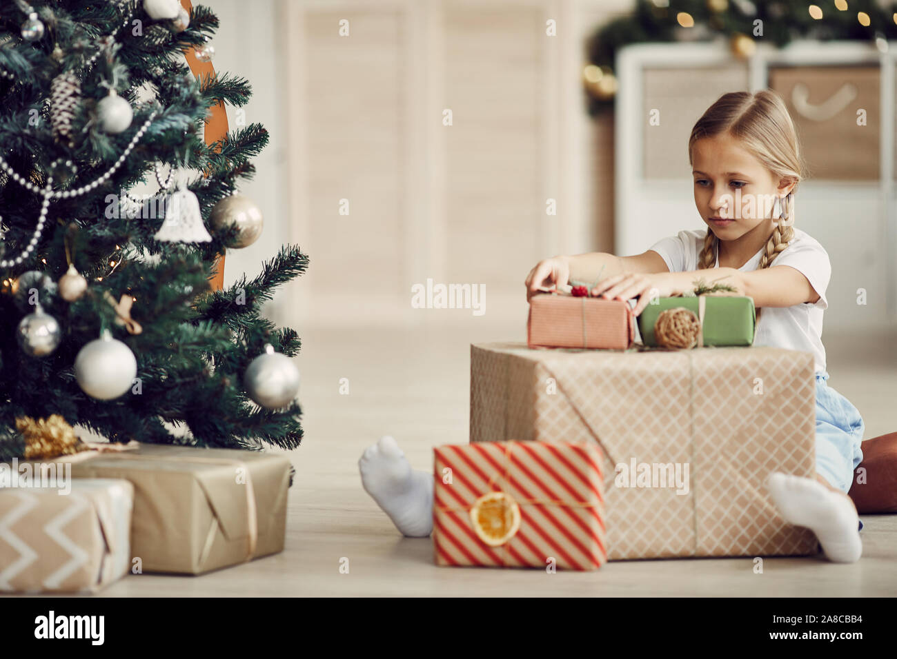Petite fille aux cheveux blonds assis sur le plancher près de l'arbre de Noël et d'examiner différentes des cadeaux de Noël à la maison Banque D'Images