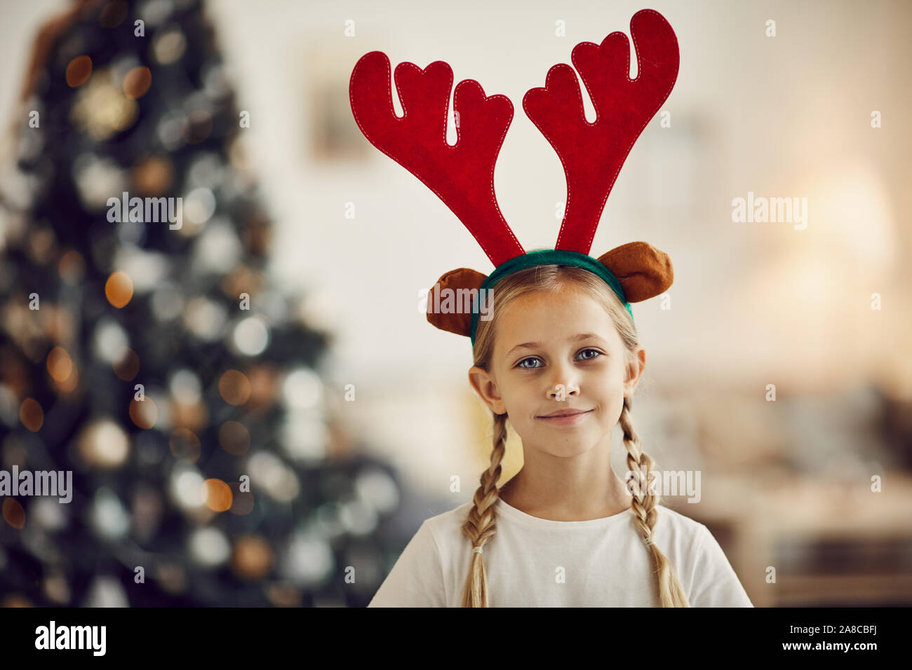 Portrait de petite fille aux cheveux blonds portant des bois de cerf et à la recherche à huis clos à la maison avec arbre de Noël dans l'arrière-plan Banque D'Images