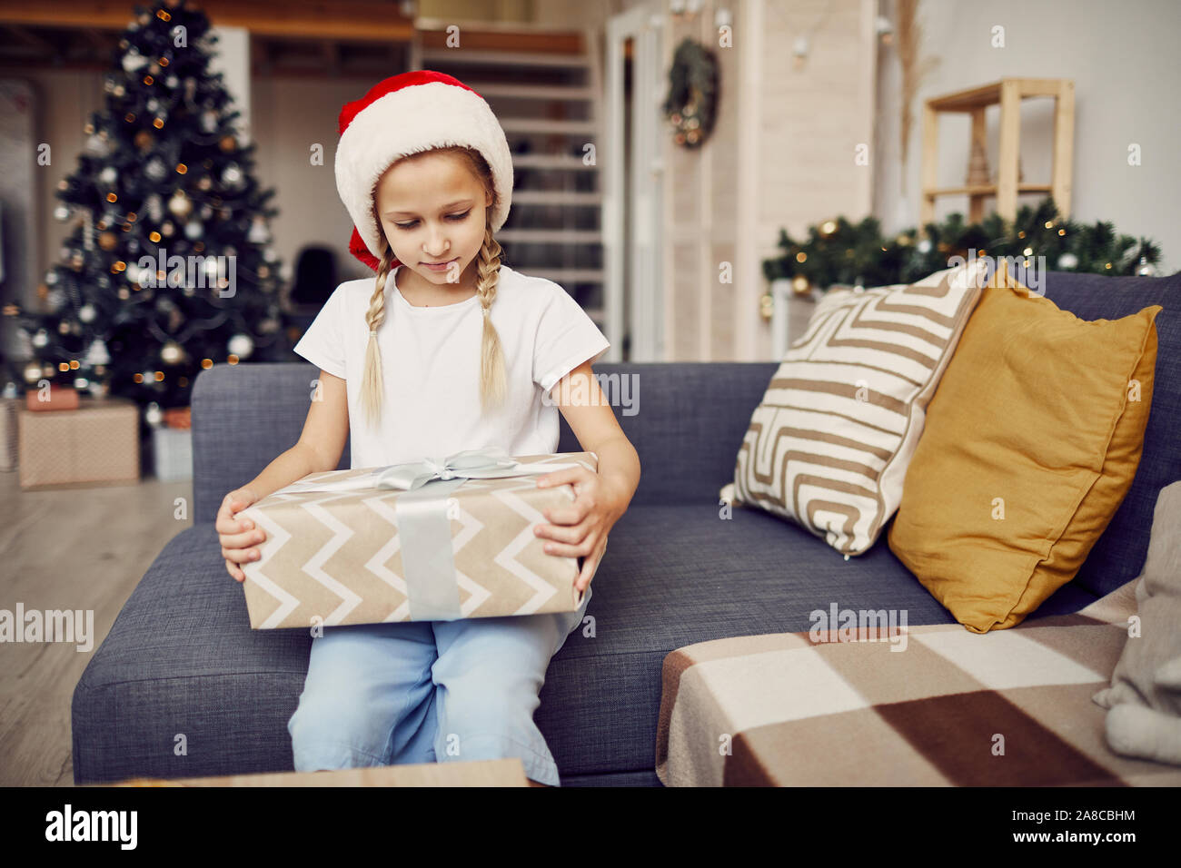 Petite fille aux cheveux blonds wearing Santa hat et assis sur un canapé avec de gros cadeau de Noël à la maison Banque D'Images