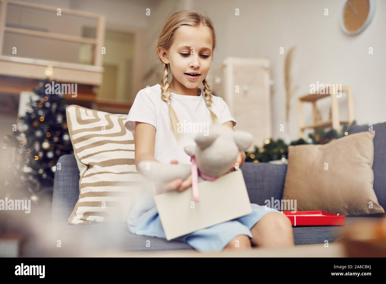 Happy little girl aux cheveux blonds à la recherche à l'ours dans ses mains pendant qu'elle l'ouverture du cadeau de Noël sur le canapé à la maison Banque D'Images