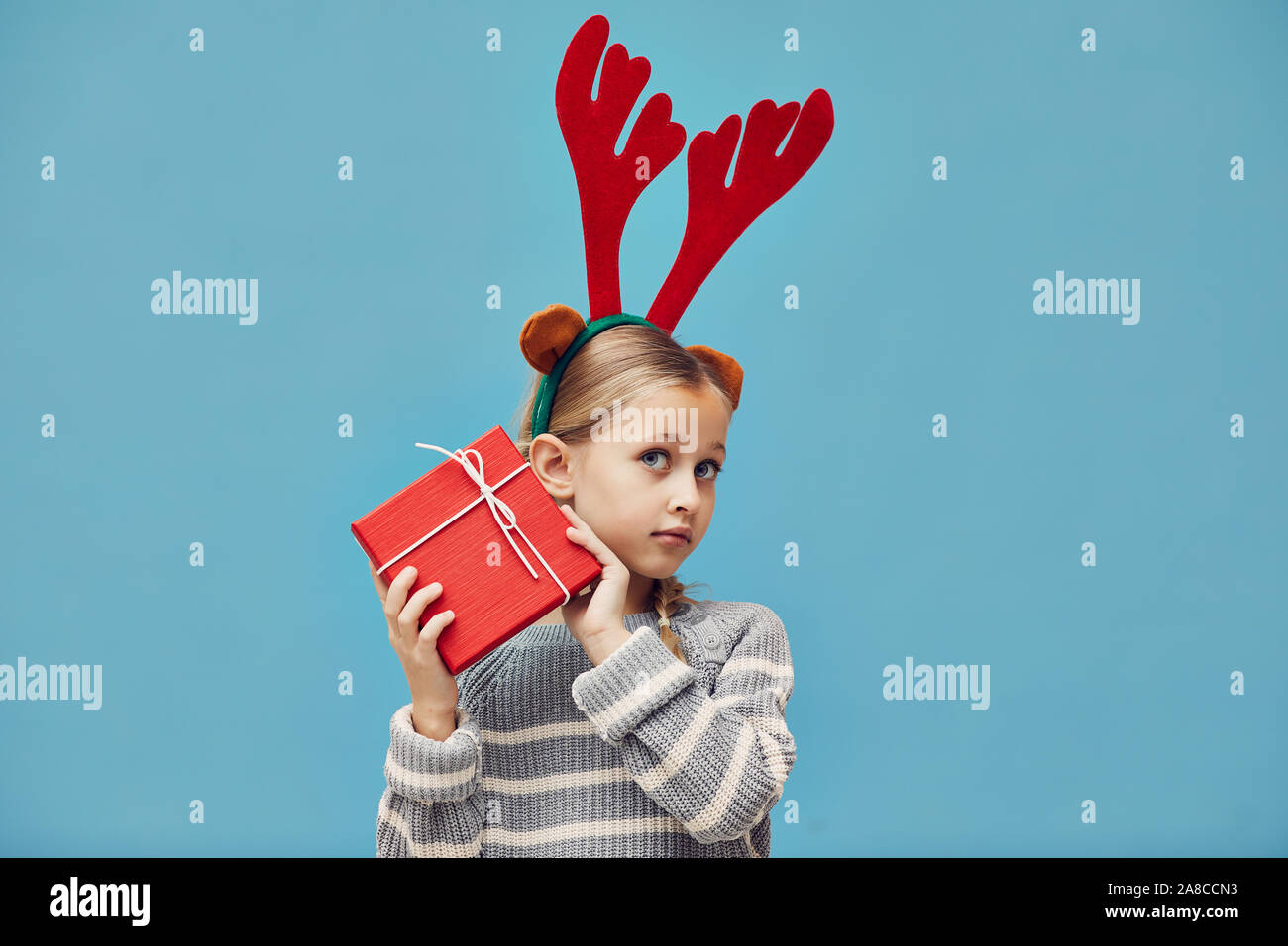 Portrait of cute little girl in deer antlers holding Christmas present dans du papier d'emballage rouge isolé sur fond bleu Banque D'Images