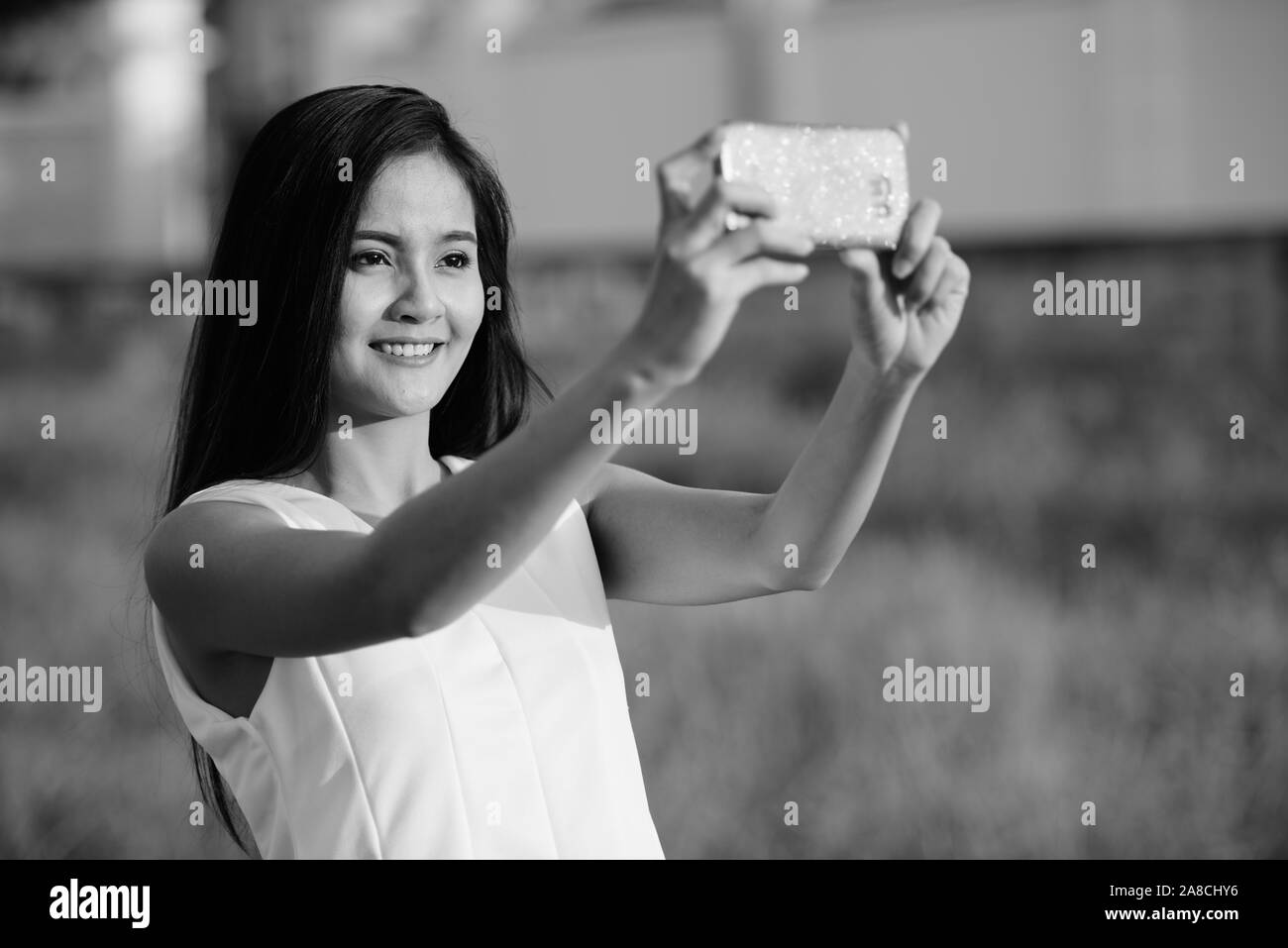 Young happy Asian woman smiling tout en tenant avec photo selfies téléphone mobile contre grass field Banque D'Images
