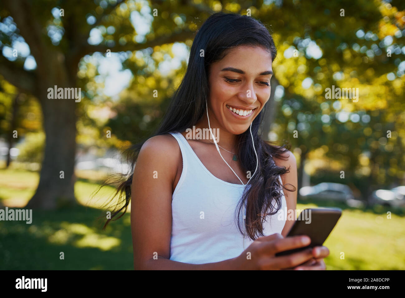 Portrait d'une jeune femme avec des écouteurs dans les oreilles à l'aide de téléphone mobile pour écouter de la musique dans le parc sous un arbre sur une journée ensoleillée Banque D'Images