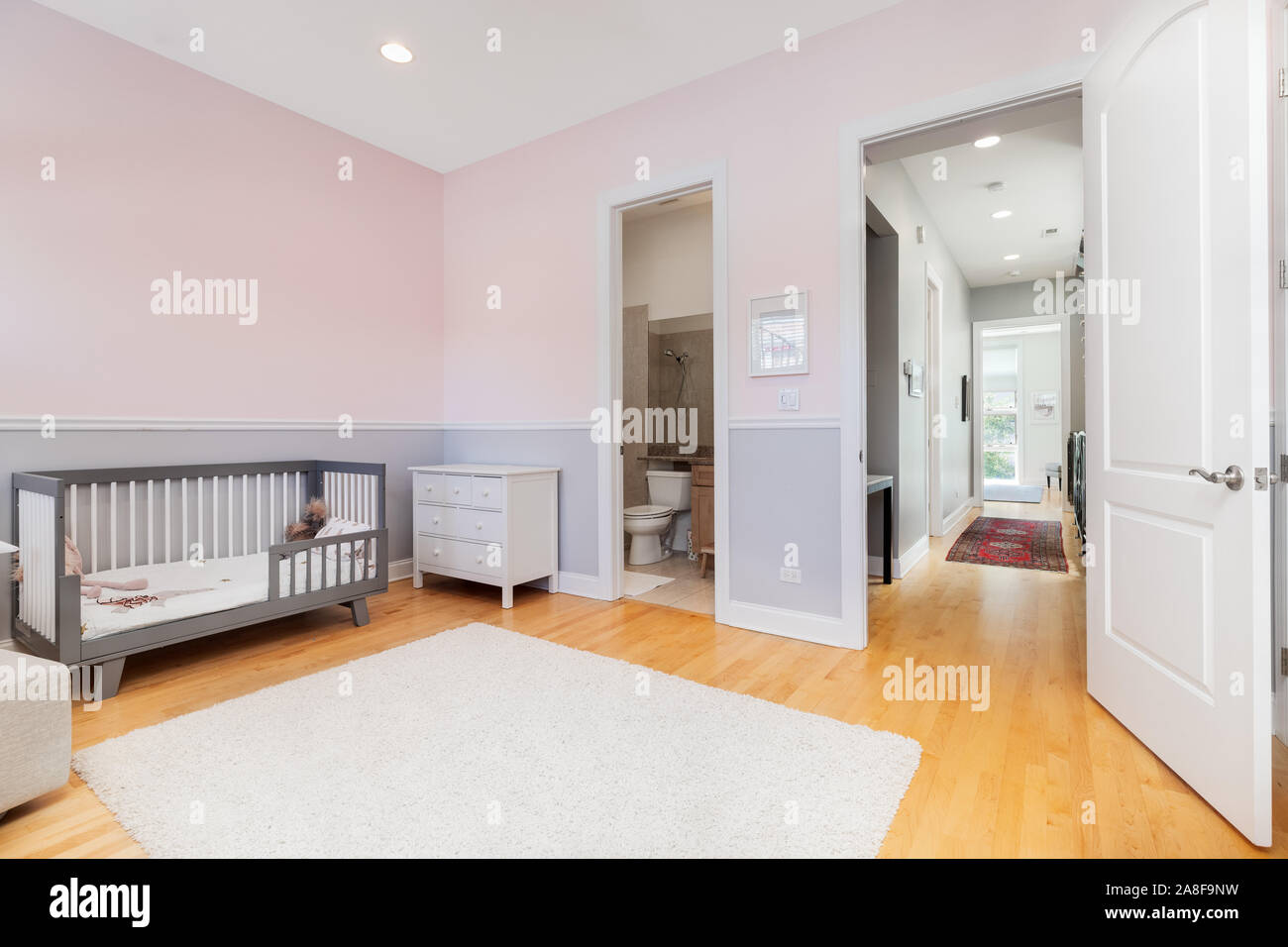 Une chambre d'enfants avec des murs blancs et violets à dans le couloir. Un lit de bébé est assis contre le mur et un tapis sur la lumière de parquet. Banque D'Images