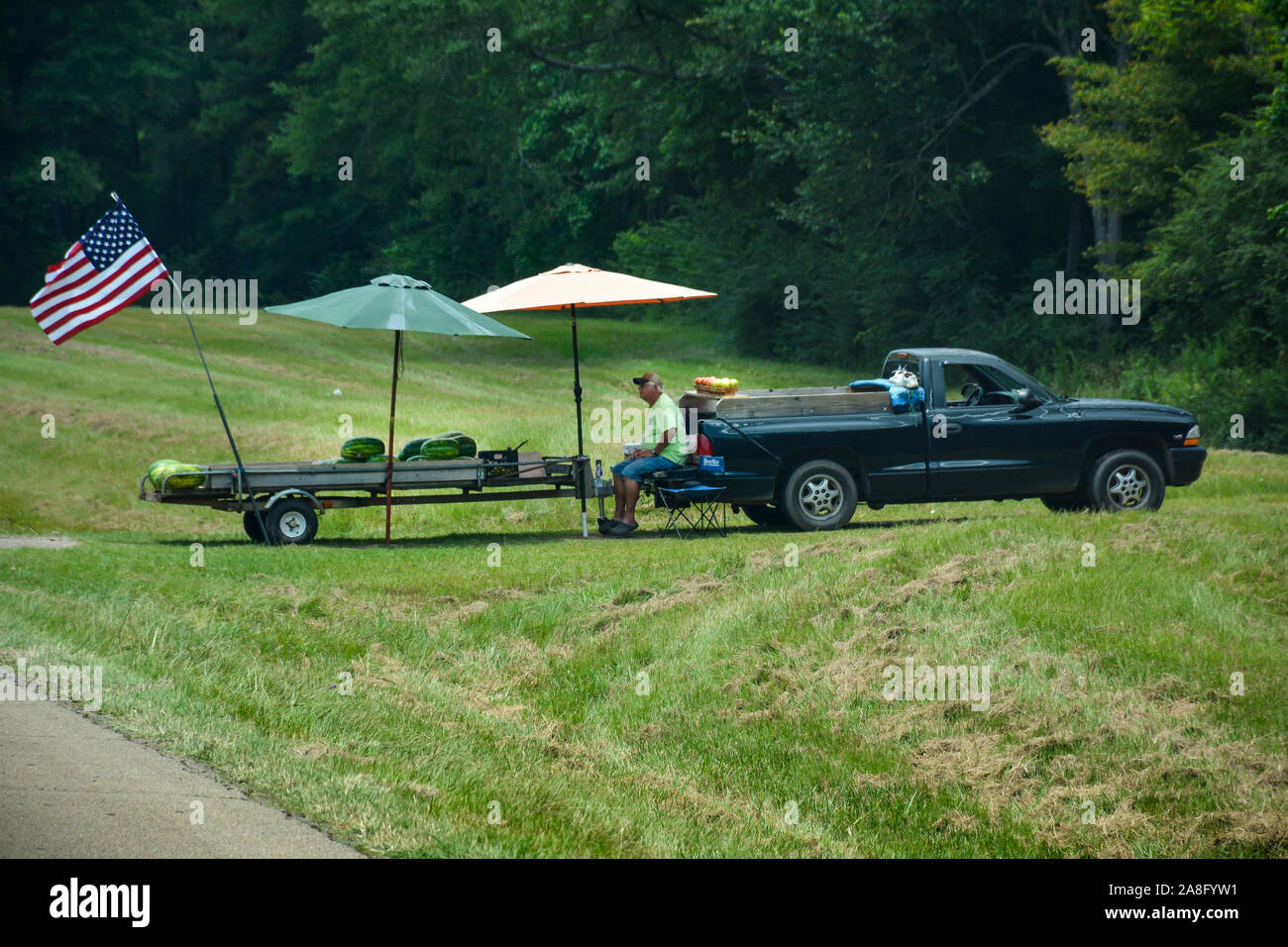 Un homme plus âgé patriotique vole le drapeau américain sur sa remorque de l'ombre des pastèques et des tomates comme un stand de fruits en bordure de route dans le sud du Mississippi Banque D'Images