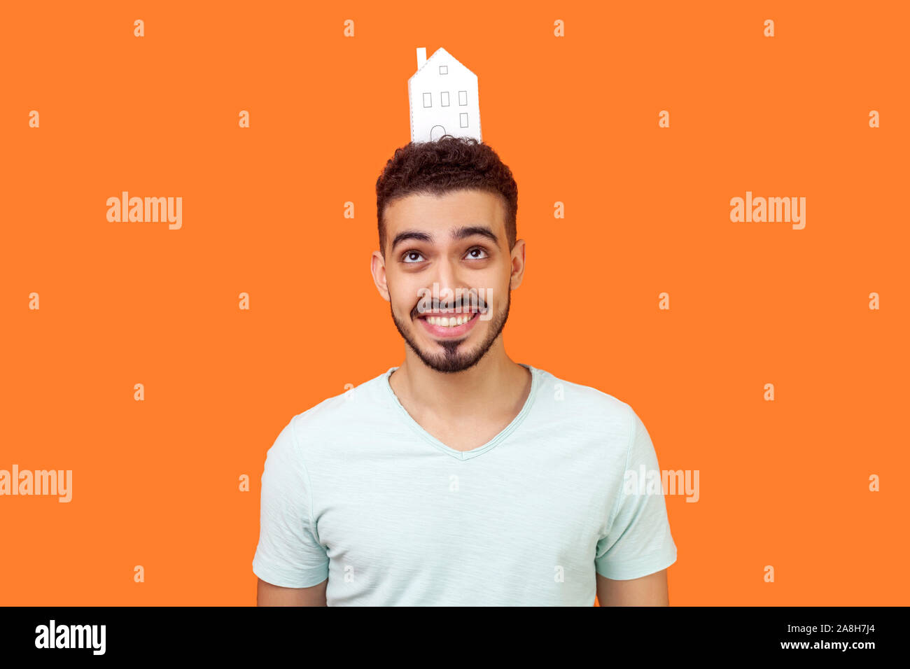 Portrait de jeune femme brune joyeuse avec l'homme à barbe blanc T-shirt standing looking up at paper house sur la tête et sourire, rêver d'accueil achat. j'ai Banque D'Images Portrait de jeune femme brune joyeuse avec l'homme à barbe blanc T-shirt standing looking up at paper house sur la tête et sourire, rêver d'accueil achat. j'ai Banque D'Images