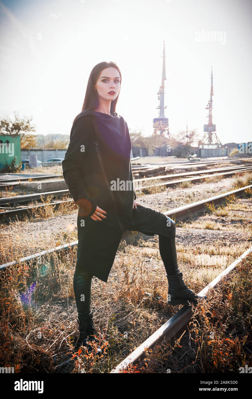 Fashion shot : portrait de la jolie fille rock (modèle informel) dans une tunique et un pantalon en cuir debout à rails (zone industrielle) Banque D'Images