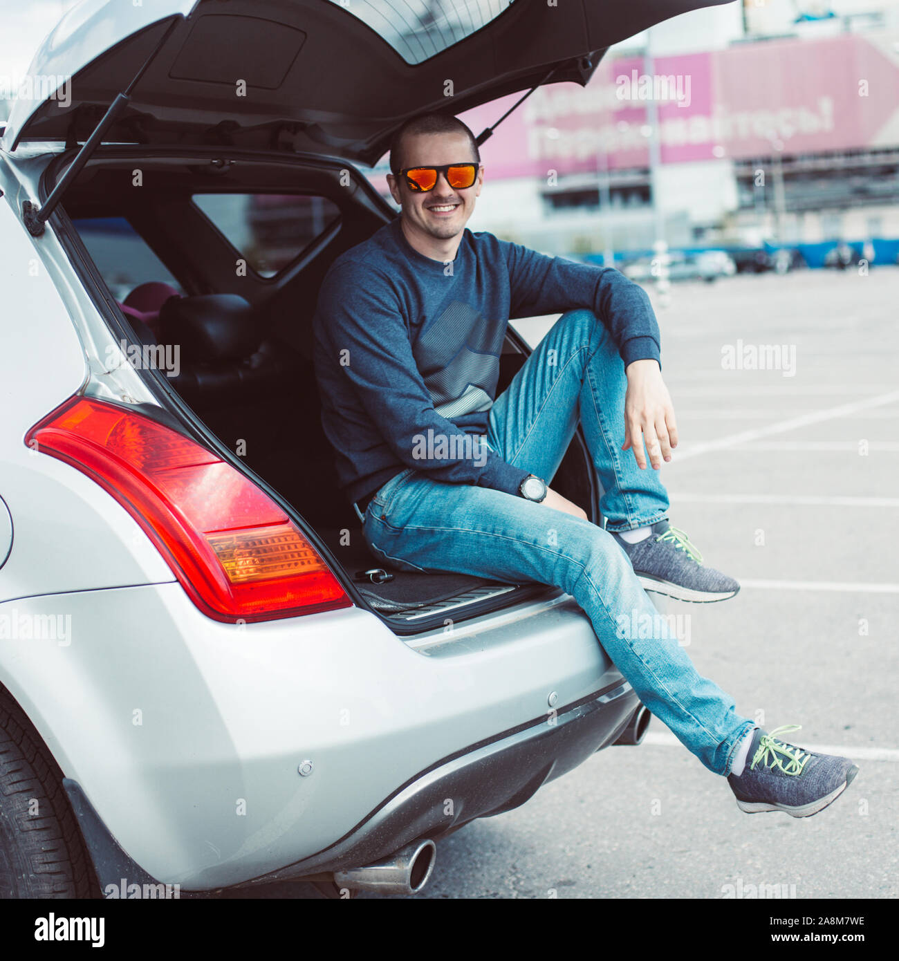 L'homme en jeans et lunettes assis dans ouvert coffre de la voiture Banque D'Images