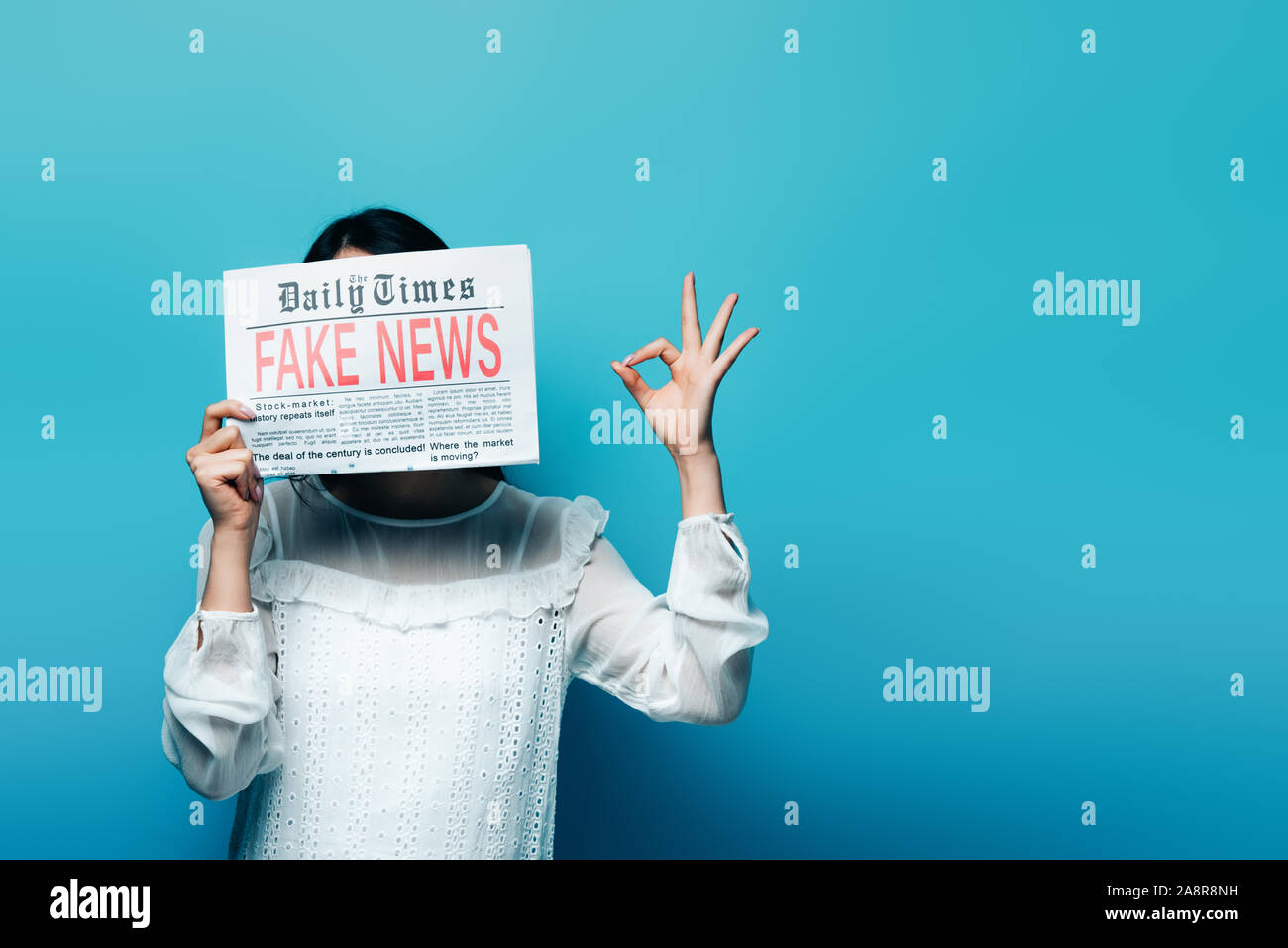 Femme en blouse blanche tenant un journal avec de fausses nouvelles et montrant ok sign on blue background Banque D'Images