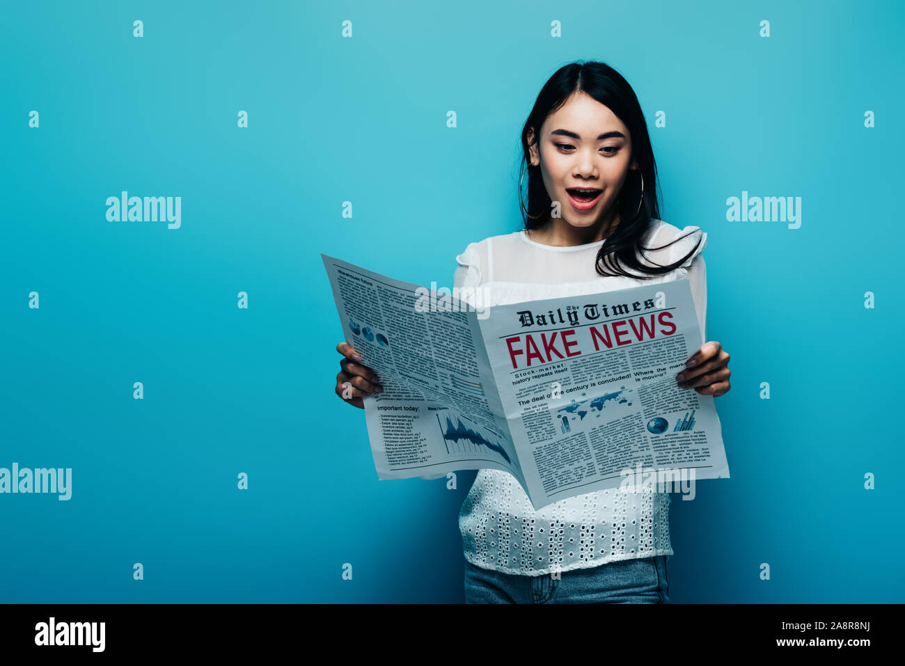Femme asiatique choqué en blouse blanche lisant le journal avec de faux news sur fond bleu Banque D'Images