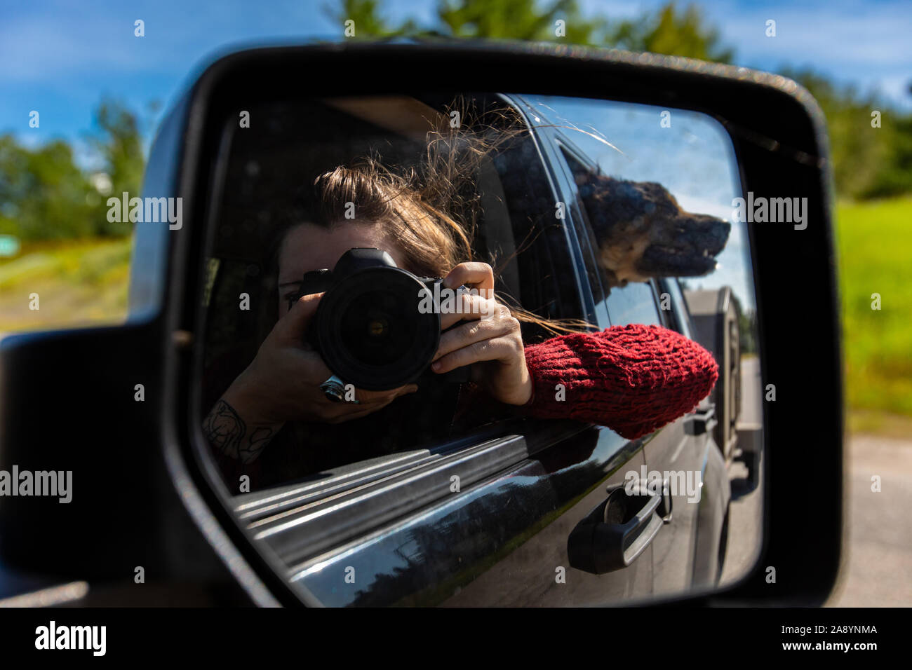 Femme photographe utilise son appareil photo reflex numérique pour faire un autoportrait à l'arrière, rétroviseur intérieur d'un SUV tout en voyageant Banque D'Images