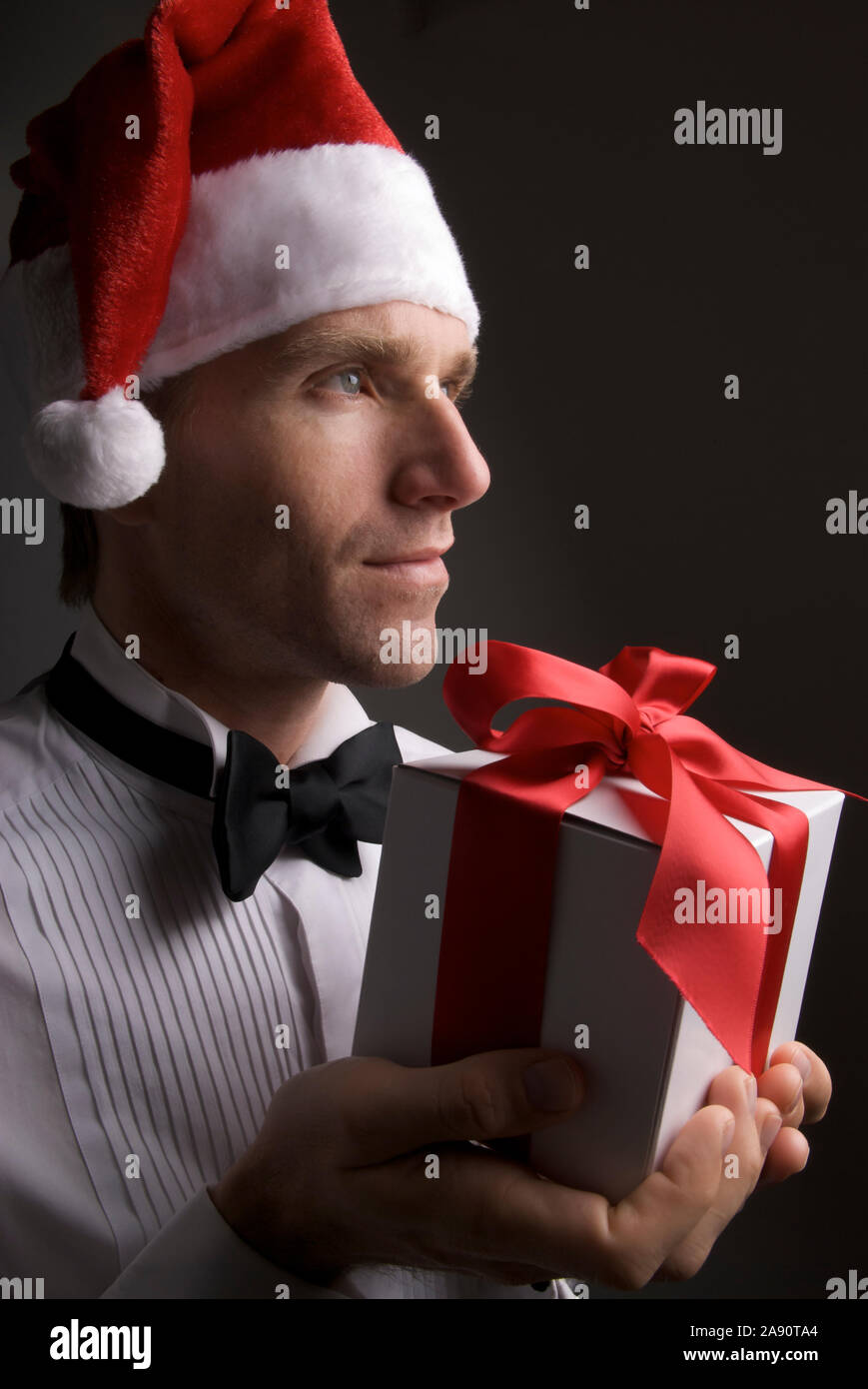 Portrait de jeune homme en smoking et cravate noire officielle Santa hat holding un cadeau avec une expression grave Banque D'Images