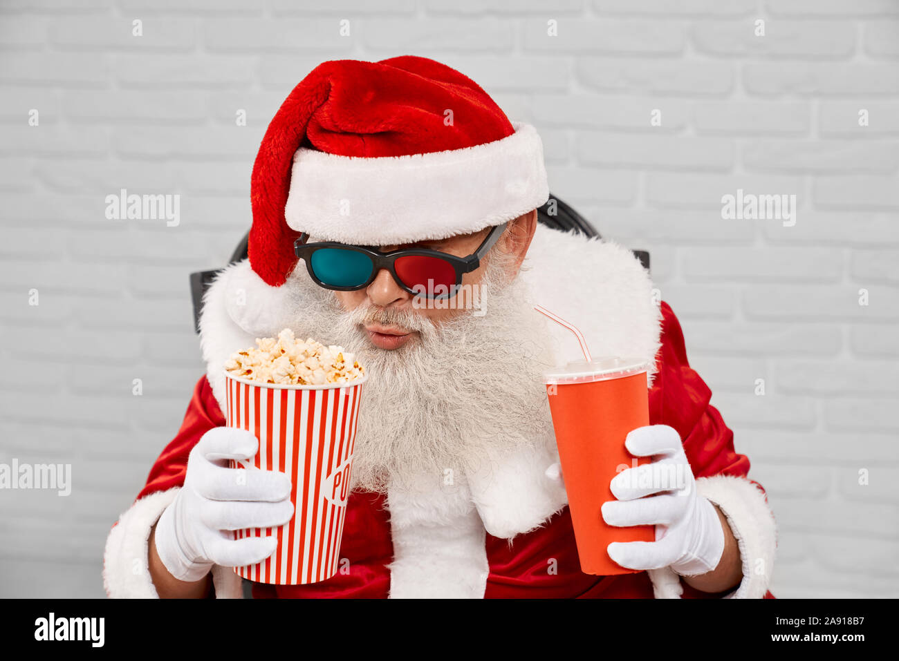 Hauts homme portant un costume traditionnel du père Noël et de lunettes 3D, assis dans un fauteuil confortable avec pop-corn et cola en white studio. Caractère noël drôle bénéficiant de temps libre. Banque D'Images