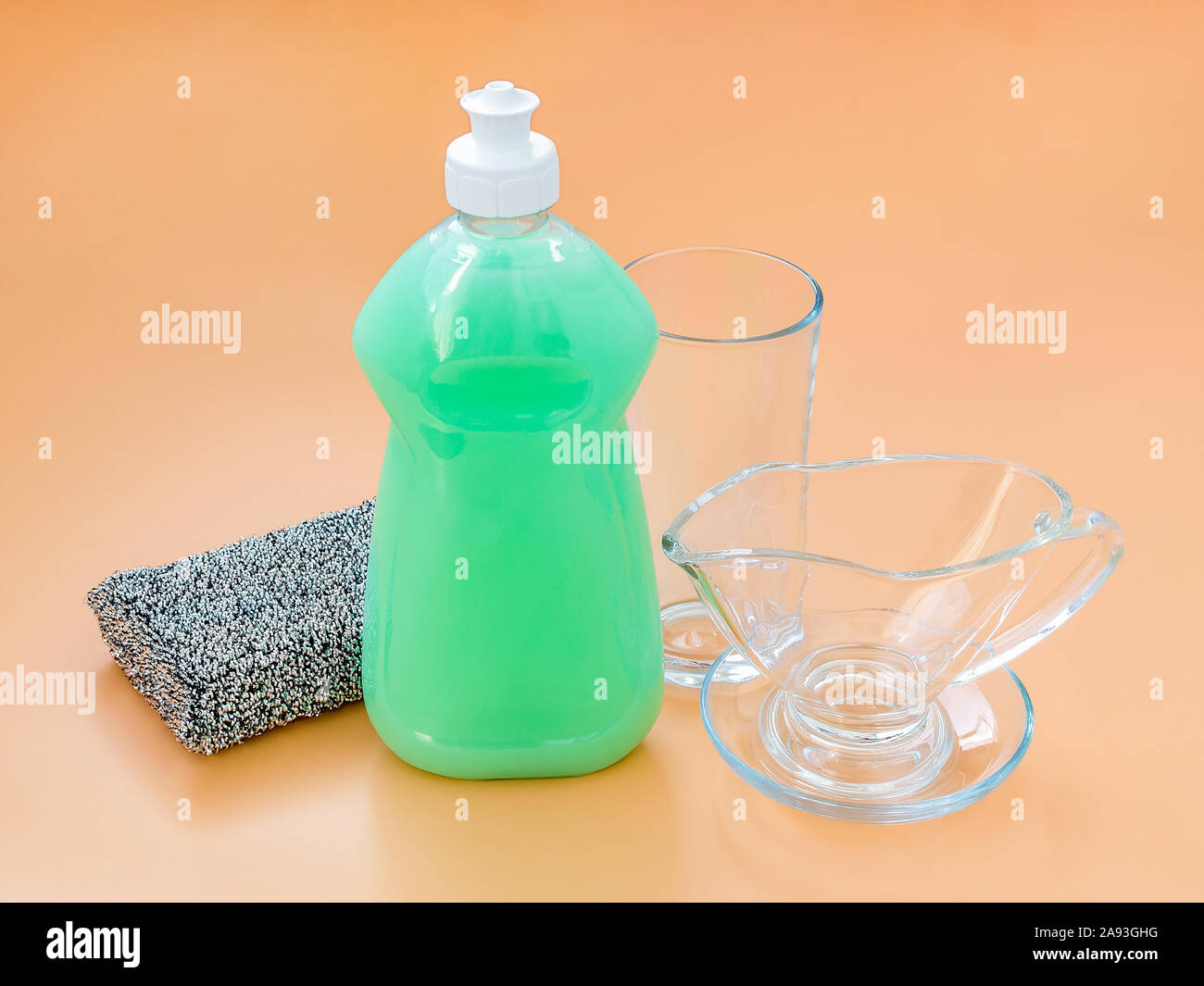 Lave-vaisselle vert liquide dans une bouteille en plastique transparent, de l'argent et la verrerie Eponge en mousse sur un fond orange pastel. Cuisine équipée d'un détergent. Banque D'Images