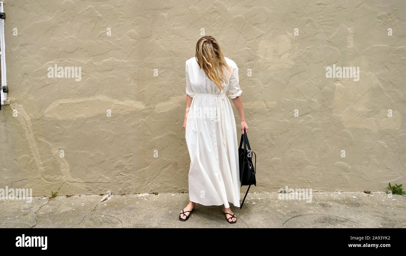 Jeune femme blonde portant une robe blanche regardant le sol devant grungy mur en stuc Banque D'Images