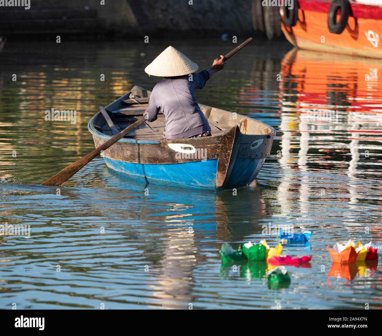Une femme de l'œil en bois bateau dans Hoi An, Vietnam avec des bateaux de papier coloré de la full moon festival à l'avant-plan. Banque D'Images