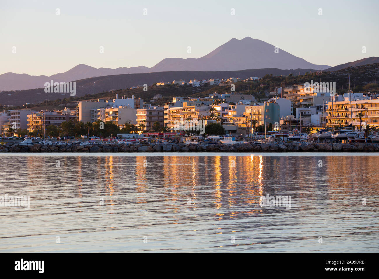 Rethymno, Crète, Grèce. Bâtiments en bord de mer se reflétant dans l'eau fixe, le lever du soleil, le Mont Psiloritis visible au-delà. Banque D'Images