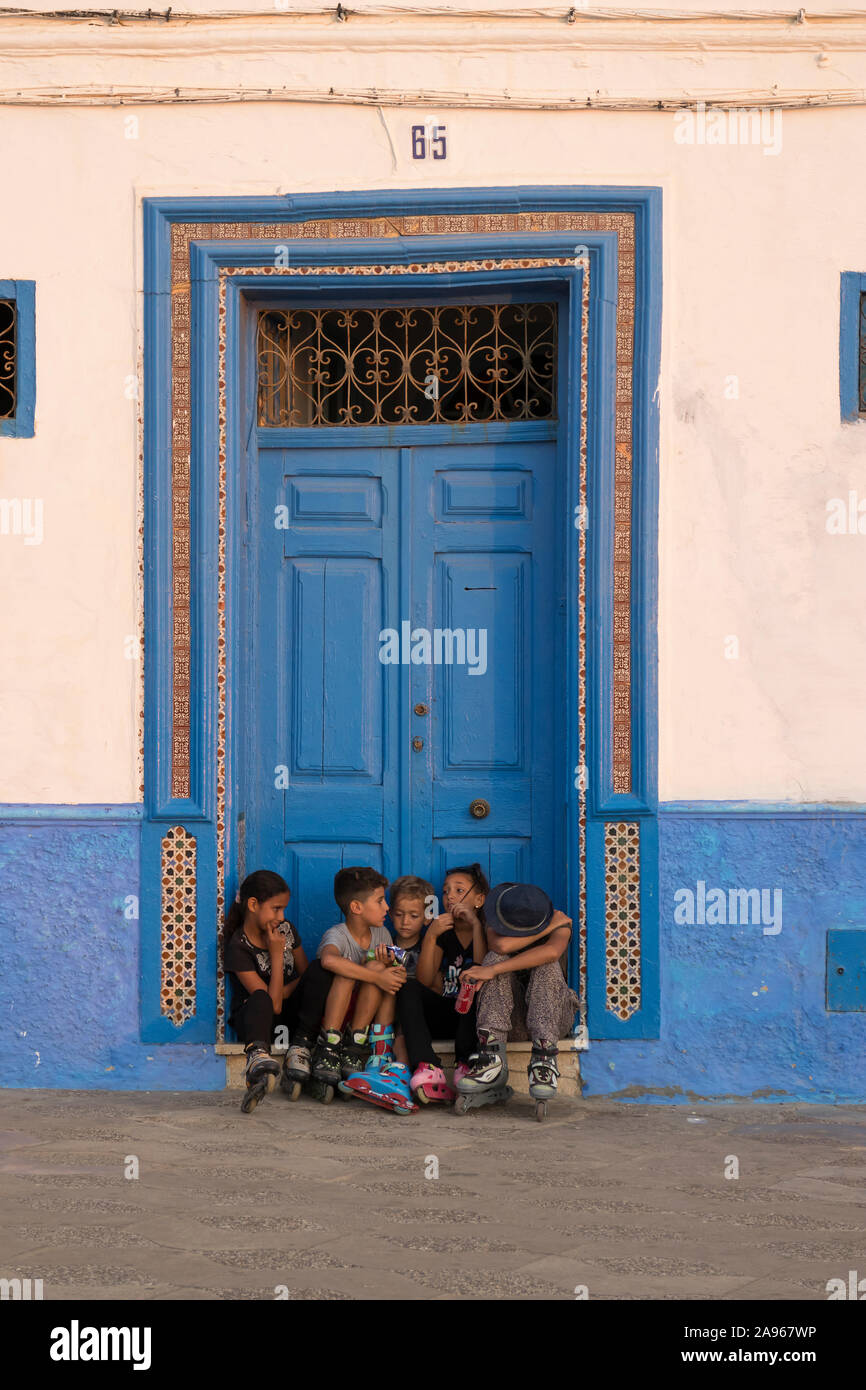 Asilah, Morocco-September 10, 2019 : des enfants assis en face d'une porte en bois peint en bleu dans la médina d'Asilah, Maroc Banque D'Images