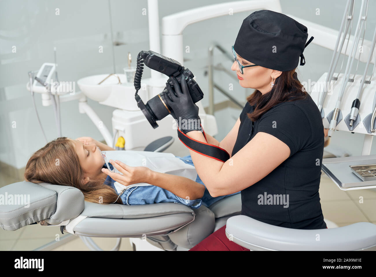 Dentiste en uniforme noir, chapeau et gants de caoutchouc médical équipement holding pour l'inspection des dents. Fille assise en fauteuil dentaire et la tenue de la bouche, de l'enrouleur à équipements à. Banque D'Images Dentiste en uniforme noir, chapeau et gants de caoutchouc médical équipement holding pour l'inspection des dents. Fille assise en fauteuil dentaire et la tenue de la bouche, de l'enrouleur à équipements à. Banque D'Images