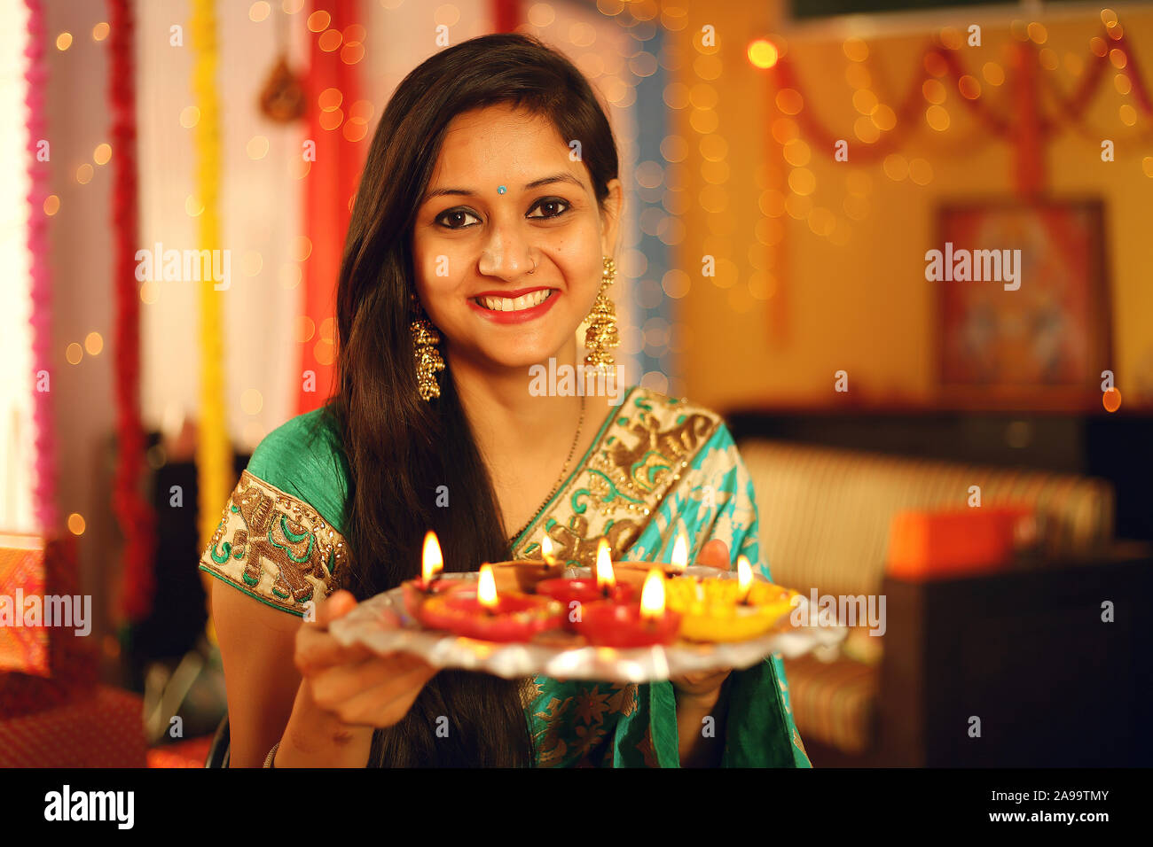 Une belle jeune femme indienne en sari traditionnel dress holding une lumière d'une lampe à huile ou diya avec en arrière-plan décoratif à l'occasion de Diwali. Banque D'Images