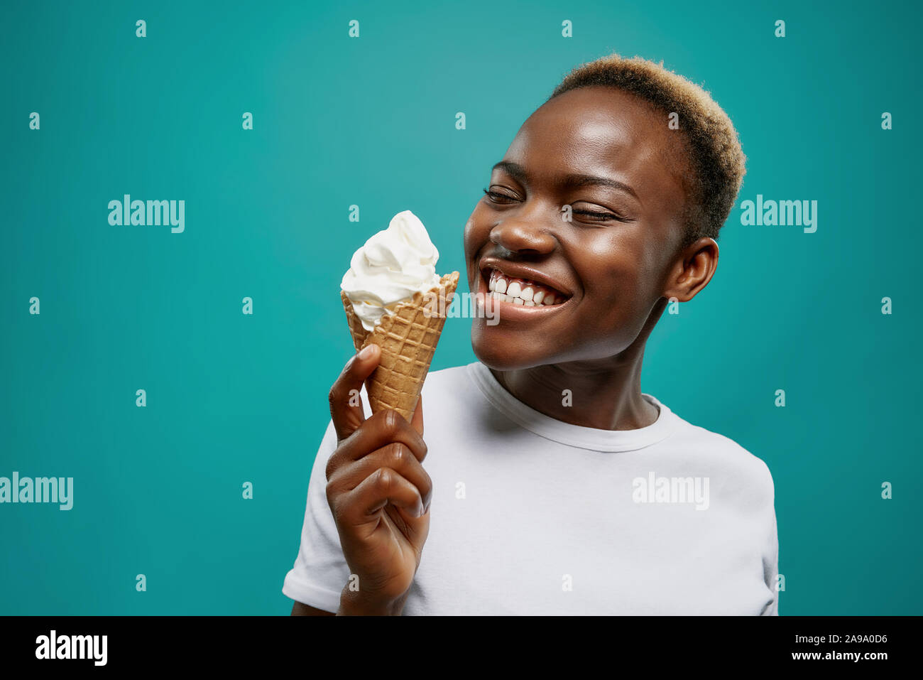 Vue latérale d'une femme joyeuse portant chemise blanche et manger de délicieux glace froide en été. Jeune fille africaine avec coupe courte à la recherche au dessert et de rire en studio. Notion de plaisir. Banque D'Images