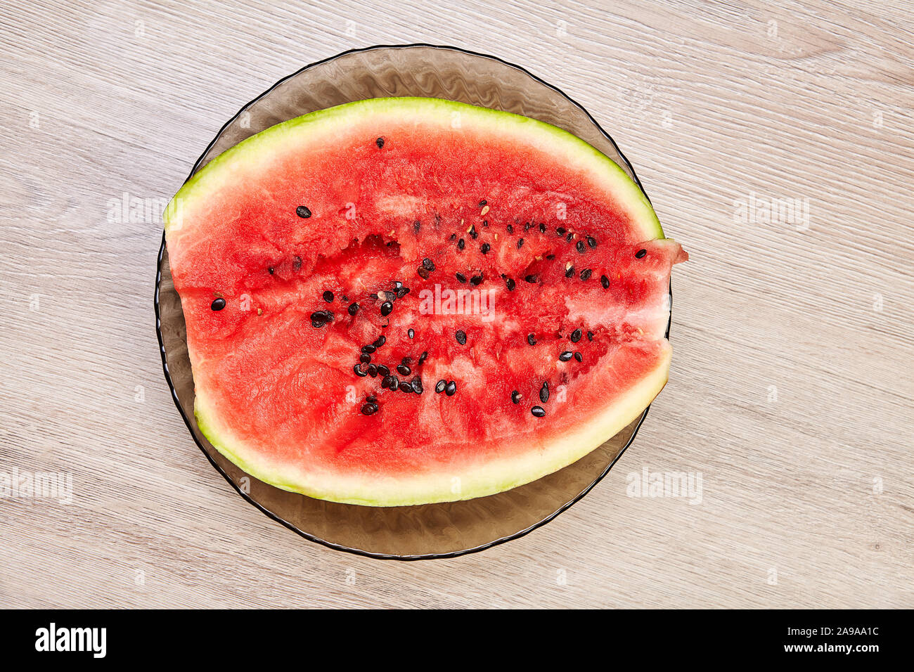 La moitié d'une pastèque rouge frais avec une coupe irrégulière se trouve sur une plaque de verre contre une table en chêne en bois blanc. Banque D'Images
