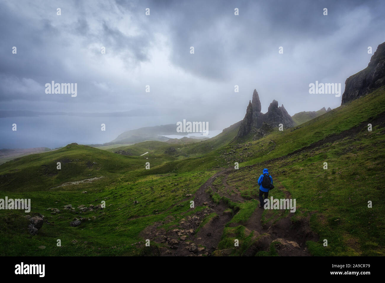 Old Man Storr. Le Storr sous un ciel nuageux dans un jour de pluie. Homme avec veste bleue de la randonnée à travers l'herbe sous la pluie. Île de Skie, en Écosse. Banque D'Images