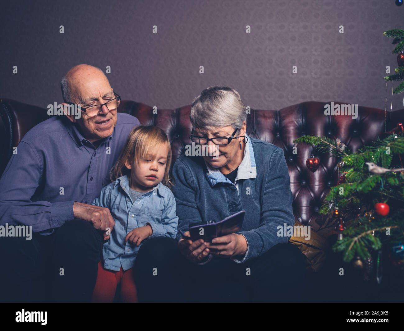 Un peu d'enfant et ses grands-parents sont assis sur un canapé en regardant un smartphone par l'arbre de Noël Banque D'Images