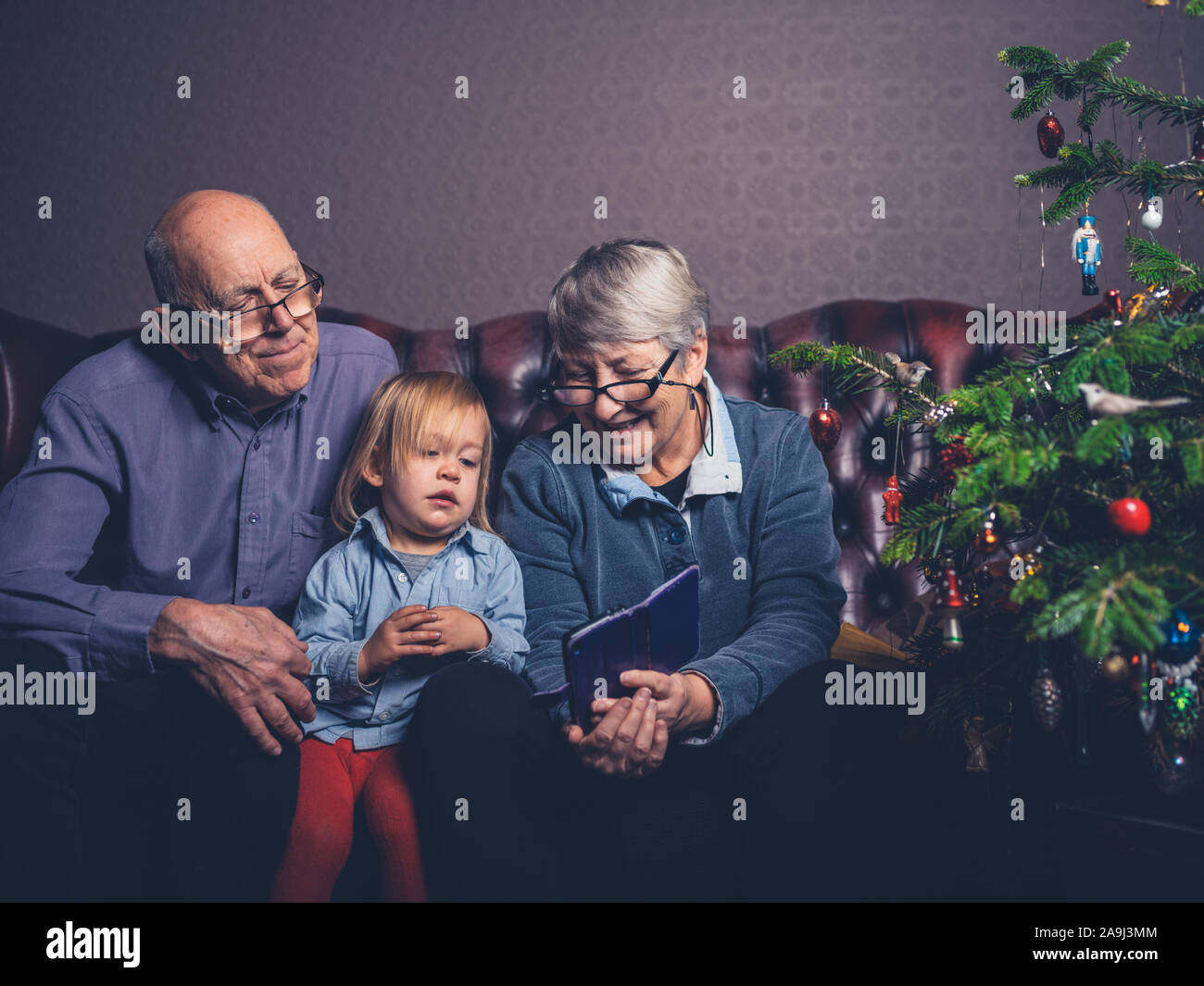 Un peu d'enfant et ses grands-parents sont assis sur un canapé en regardant un smartphone par l'arbre de Noël Banque D'Images