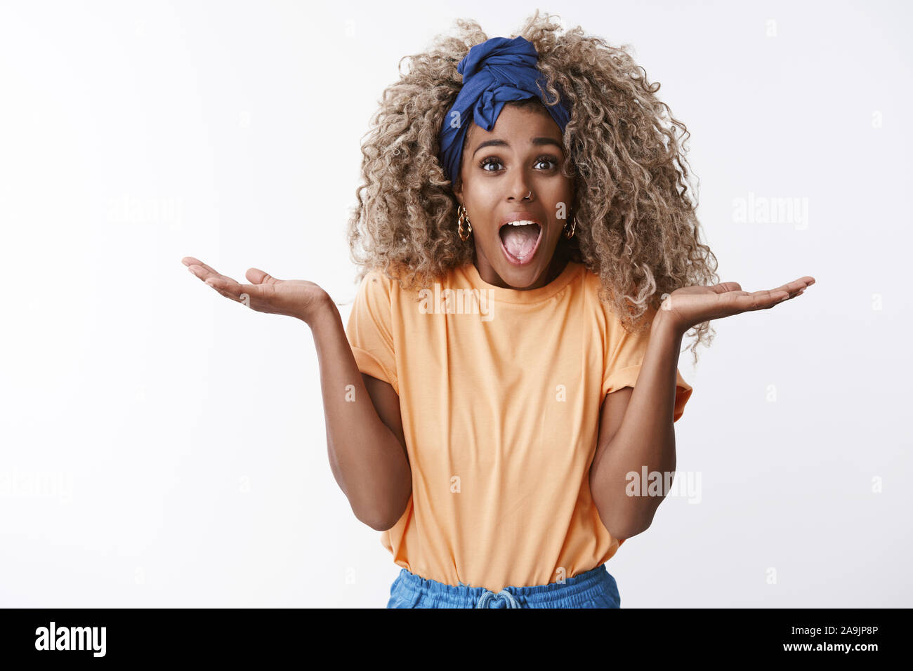 Bien charismatiques africains-américains curly blonde woman in stylish t-shirt orange, bandeau, cris amusés, lever les mains sur le côté et un impressionné Banque D'Images